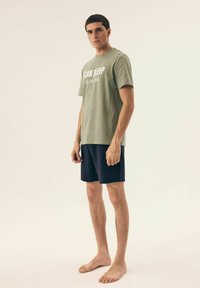 Oliwkowa bawełniana koszulka z białym napisem, połączona z granatowymi szortami. Luźny krój, krótkie rękawy i prosty design.