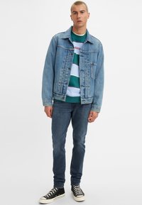Denim jacket with a light blue hue, paired with a green striped sweater and dark jeans. Black sneakers complete the look.