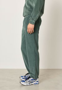 Žepri zeleni kordurojski hlače z elastičnim pasom, dopolnjene s srebrno-modrimi športnimi čevlji. Stranski pogled prikazuje sproščen kroj.
