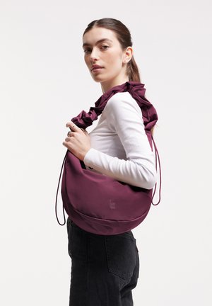 Bolso de hombro morado con correa fruncida, hecho de material suave, con forma redonda y una marca discreta en la parte frontal.
