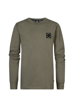 Jugendlicher trägt ein olivgrünes Langarmshirt mit kleinem Brustlogo, blaue Jeans und schwarz-weiße Sneakers, steht mit einer Hand in der Tasche.