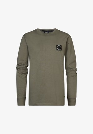 Jeune portant une chemise à manches longues vert olive avec un petit logo sur la poitrine, jean bleu et baskets noir et blanc, debout avec une main dans la poche.