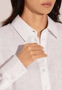 Weißes Leinenhemd mit strukturiertem Kragen, Knopfleiste und Manschettendetails. Der Stoff hat ein strukturiertes, atmungsaktives Aussehen. Handgenähte Anpassungen sind sichtbar.