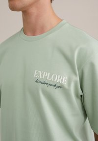 Világoszöld pamut póló kerek nyakkivágással, fehér nyomott felirattal, amely az "EXPLORE" és a "let nature guide you" szöveget eltérő stílusokban tartalmazza.
