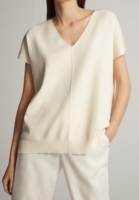 Kermanvalkoinen V-kaula-aukollinen toppi, jossa on lyhyet hihat, pehmeästä kankaasta valmistettu, rento istuvuus ja resorireunus. Yhdistetty vaaleisiin, vastaaviin housuihin.