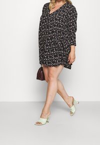 Vestido negro de flores con mangas largas y escote en V; presenta pequeñas flores rosas y verdes. Combinado con mules de tacón verde claro y un bolso marrón.