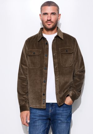 Mand med kort hår og skæg iført en brun kordjakke over en hvid skjorte og blå jeans, stående med den ene hånd i lommen.