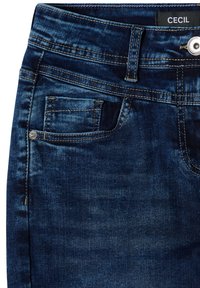 Jean en denim bleu foncé en gros plan montrant une poche avant, une petite poche à monnaie, un bouton, une boucle de ceinture et une étiquette noire avec le texte "CECIL".