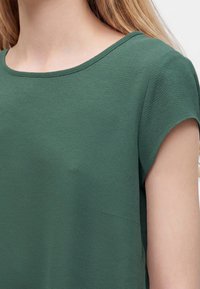 Gros plan d'une personne portant un haut vert texturé à manches courtes et col rond avec des longs cheveux blonds visibles.