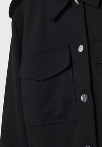 Chemise noire à manches longues avec deux poches poitrine à rabat et boutons-pression argentés brillants visibles sur le devant.