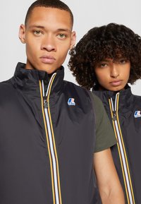 To personer har på seg svarte, ermeløse vester med høy krage og flerfargede striper på glidelåsen. Vestene har en logo på brystet.