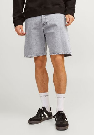 Mężczyzna w jasnoniebieskich dżinsowych szortach, czarno-białych tenisówkach, białych skarpetkach do kostek i czarnej bluzie z długim rękawem, stojący na jednolitym tle.