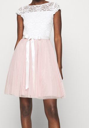 Femme portant une robe courte avec un haut en dentelle blanche, des manches courtes et une jupe en tulle rose nouée avec une ceinture ruban en satin blanc.
