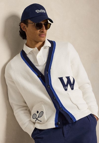 Άνδρας που φοράει γυαλιά ηλίου, σκούφο Wimbledon σε ναυτικό μπλε, λευκή ζακέτα με μπλε περιθώριο και κέντημα με ρακέτα του τένις, λευκό πόλο και ναυτικό μπλε παντελόνι.