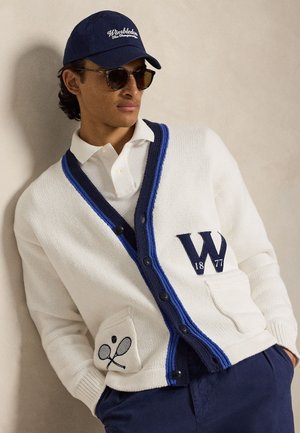 Άνδρας που φοράει γυαλιά ηλίου, σκούφο Wimbledon σε ναυτικό μπλε, λευκή ζακέτα με μπλε περιθώριο και κέντημα με ρακέτα του τένις, λευκό πόλο και ναυτικό μπλε παντελόνι.