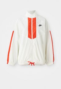 Fehér szélálló dzseki piros függőleges csíkokkal és húzózsinóros aljjal. Álló gallérral és elől cipzárral rendelkezik. A mellkason Nike logó található.
