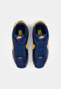 Nike tenisice imaju pretežno mornarsko plavu gornju stranu, žute akcentne detalje i teksturiranu završnu obradu. Uključuju klasično vezivanje i klasičan dizajn.