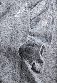 Tejido de punto gris con lentejuelas dispersas, que presenta una textura suave y bordes ondulados. Un sutil patrón jaspeado añade profundidad al diseño.