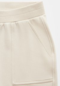 Beige Jogginghose mit geripptem, elastischem Bund, zwei Seitentaschen und abgesteppten Details an Vorderseite und Taschen. Weicher, strukturierter Stoff.