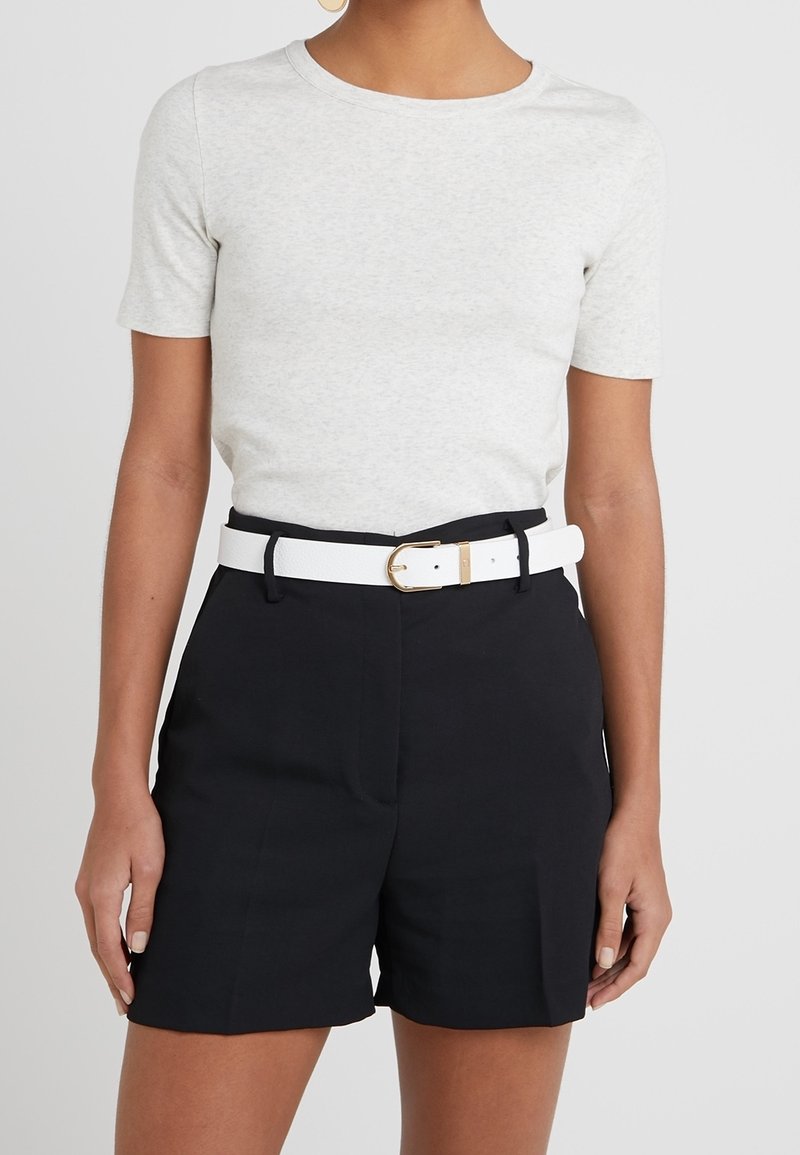 Femme portant un t-shirt gris clair à manches courtes rentré dans un short noir taille haute avec une ceinture blanche et une boucle dorée.