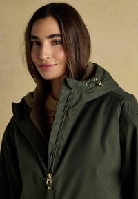 Olivengrøn jakke med en blød, brun pelsforet krave; har en frontlynlås, guldfarvet metaldele og branding langs lynlåskanten.