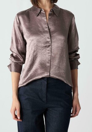 Kobieta ubrana w długą, marszczoną, satynową bluzkę w kolorze mauve z ostrym kołnierzykiem, zestawioną z ciemnoniebieskimi spodniami z wysokim stanem.