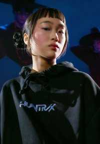 Čierna mikina s kapucňou so sťahovacou šnúrkou, s logom "HUNTRX" v reflexnom modrom písme. Hladká textúra, priliehavý strih a mäkká tkanina.