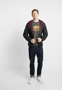 Chaqueta acolchada negra con capucha burdeos, llevada sobre una camiseta gris con logo. Combinada con jeans oscuros y zapatillas blancas.