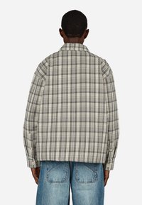 Beige és fekete szövetből készült nagy méretű kockás ing. Gallérral és gombbal záródó mandzsettákkal rendelkezik. A hátoldalon eltérő kockás minta látható.