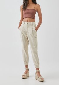 Beige tapered broek met elastische boorden, voorzien van zijzakken en een kettingaccent. Gecombineerd met een bruin geribbeld crop top en platform sandalen.