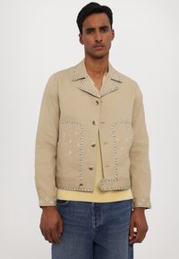 Giacca cropped beige con cuciture in contrasto blu e bianche, chiusura con bottoni, due tasche frontali e ricami dettagliati sui bordi.