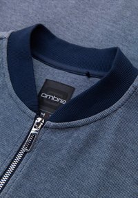 Chaqueta azul de cremallera con un tejido texturizado, cuello de punto azul marino y detalle de cremallera. La etiqueta dice "ombre" y es de talla "M".