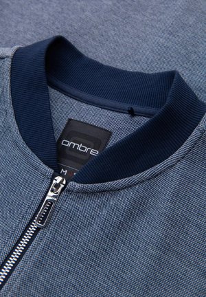 Blauwe ritsjack met een gestructureerde stof, een geribde marineblauwe kraag en een ritsaccent. Het label leest "ombre" en de maat is "M."