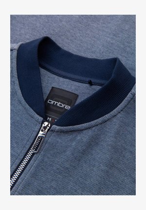 Blå zip-up jacka med strukturerat tyg, ribbad marinblå krage och dragkedjeaccent. Märket visar "ombre", i storlek "M."