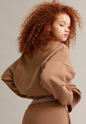 Jeune personne aux cheveux roux bouclés et volumineux, portant un sweat-shirt marron ample et un pantalon assorti, regardant par-dessus son épaule.