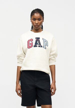 Frau mit geflochtenem Haar, die ein cremefarbenes Sweatshirt mit bunten gemusterten "GAP"-Buchstaben und schwarze kniehohe Shorts trägt, steht vor einem weißen Hintergrund.