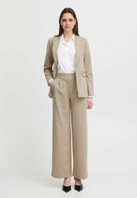 Beige kostym med en skräddarsydd blazer och vida byxor, lager på lager över en vit skjorta med knappar. Svarta spetstövlar med pekad tå fulländar outfiten.