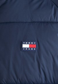Veste isolée bleu marine avec une patch rectangulaire en tissu portant "TOMMY JEANS" et un logo avec des éléments rouges, blancs et bleus. Texture lisse.
