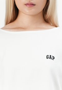 Detailní záběr na osobu s bílou košilí a černým logem "GAP" na levé hrudi, s dlouhými blond vlasy v záběru.