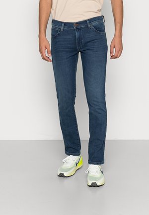 Vaqueros de denim azul oscuro con un corte slim, con cinco bolsillos, costuras sutiles y un cierre de botón clásico. Combinados con zapatillas ligeras.