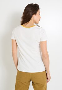 Camiseta blanca de manga corta con un cuello acanalado amarillo, vista desde la parte trasera. La tela parece suave con una textura lisa.