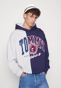 Hetted genser delt i grå og marineblå, med "TOMMY"-logo og globusgrafikk i rød og hvit. Fleece-textur, kengurulomme, avslappet passform.