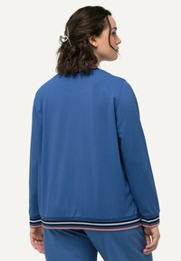 Giacca sportiva blu, tessuto leggero, vestibilità rilassata, polsini a coste con strisce multicolori, vista posteriore uniforme, design minimalista.