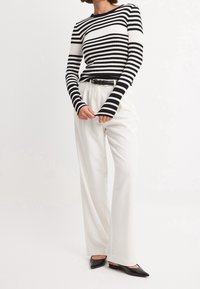 Personne portant un haut à manches longues rayé noir et blanc, un pantalon blanc taille haute à jambes larges avec une ceinture noire, et des ballerines noires à bouts pointus, sur fond blanc.