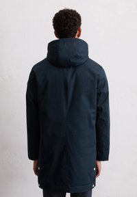 Man met kort krullend haar, gekleed in een marineblauwe lange hooded jacket, kijkend naar een eenvoudige witte muur, met ontspannen handen langs zijn zij.