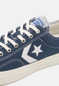 Blå canvas sneakers med vit gummisula. Har vitt stjärnlogotyp och kontrasterande sömmar. Bomullsnöre och vadderad krage.