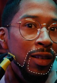 Electric razor with a yellow handle trims facial hair on a man’s jawline. He has glasses and short hair, with well-groomed facial hair.