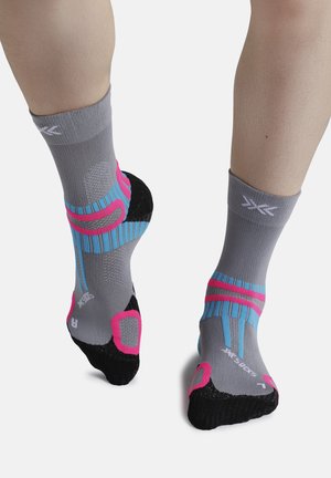 Beine, die graue Sportsocken mit pinken, blauen und schwarzen Polsterelementen tragen, aus Rücken- und Vorderansicht gezeigt.