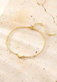 Pulsera de cadena de oro con un colgante en forma de corazón, que presenta un acabado pulido, un diseño de eslabones y un cierre ajustable, todo sobre una superficie texturizada.