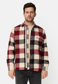Rutet flannelskjorte i røde, sorte og cremefarver. Lavet af blødt stof med knapper foran og to brystlommer.